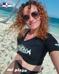 Frau mit roten Haaren und Sonnenbrille am Strand mit Meer im Hintergrund und schwarzes Hi-Fish T-Shirt Mujer pelirroja con gafas de sol en la playa con mar de fondo y camiseta negra Hi-Fish